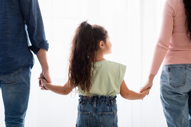 Concetto di sostegno familiare. Vista posteriore della bambina che si tiene per mano con mamma e papà, in piedi vicino alla finestra di casa, primo piano di genitori e figli di sesso femminile che si legano insieme, immagine ritagliata