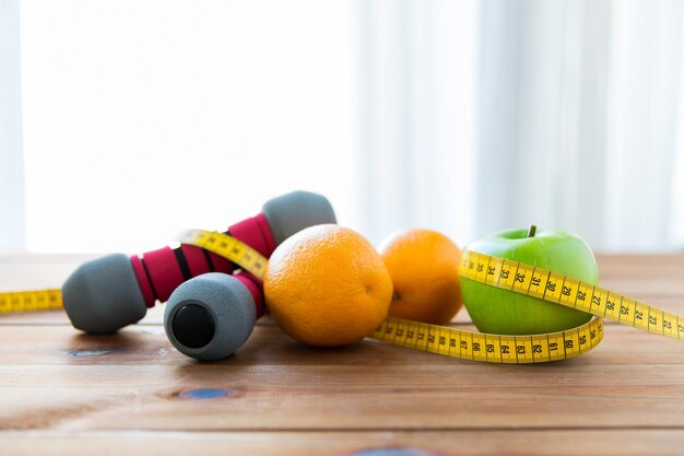concetto di sport, fitness, dieta e oggetti - primo piano di manubri e mela verde con arance avvolte da nastro di misurazione su tavolo di legno
