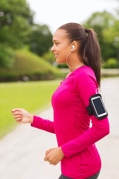 concetto di sport, fitness, salute, tecnologia e persone - giovane donna afroamericana sorridente che corre con smartphone e auricolari all'aperto