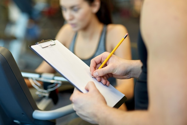 concetto di sport, fitness, stile di vita, tecnologia e persone - primo piano delle mani dell'allenatore con la scrittura negli appunti e la donna che si allena sulla cyclette in palestra