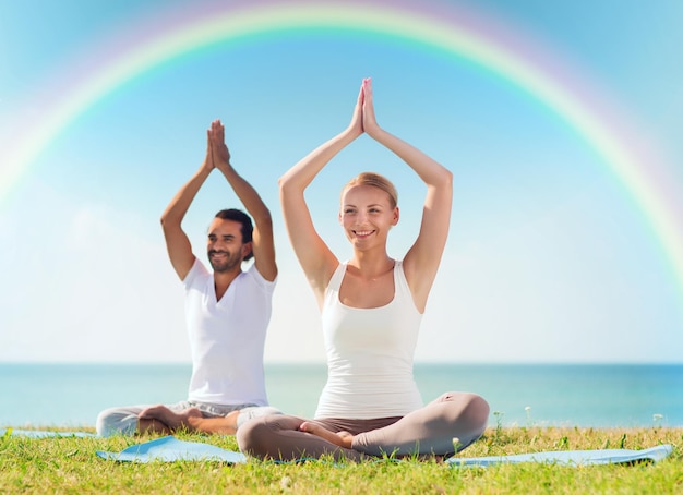 concetto di sport, fitness, yoga e persone - coppia sorridente che medita e si siede su stuoie con le mani alzate sul mare e arcobaleno sullo sfondo del cielo blu