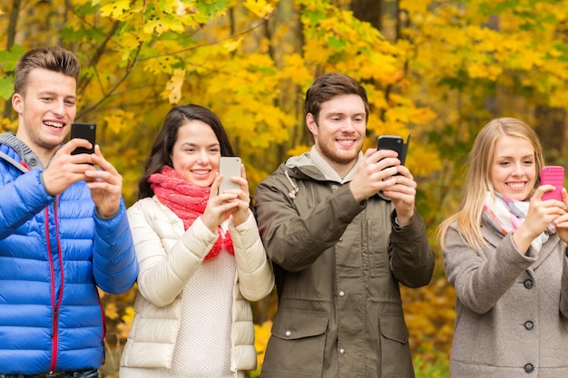 concetto di stagione, persone, tecnologia e amicizia - gruppo di amici sorridenti con smartphone che scattano foto nel parco autunnale