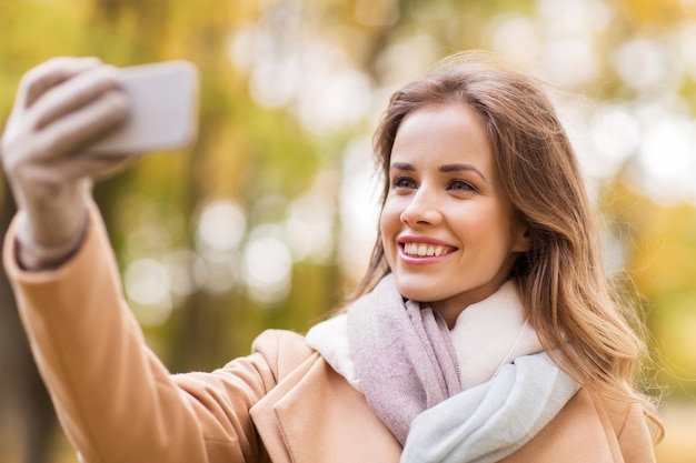 concetto di stagione, tecnologia e persone - bella giovane donna felice che prende selfie con lo smartphone nel parco autunnale
