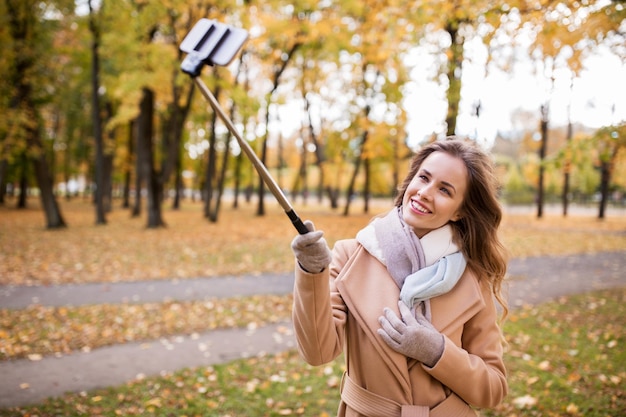 concetto di stagione, tecnologia e persone - bella giovane donna felice che scatta foto con il bastone selfie dello smartphone nel parco autunnale