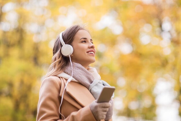 concetto di stagione, tecnologia e persone - bella giovane donna felice con le cuffie che ascolta musica sullo smartphone che cammina nel parco autunnale