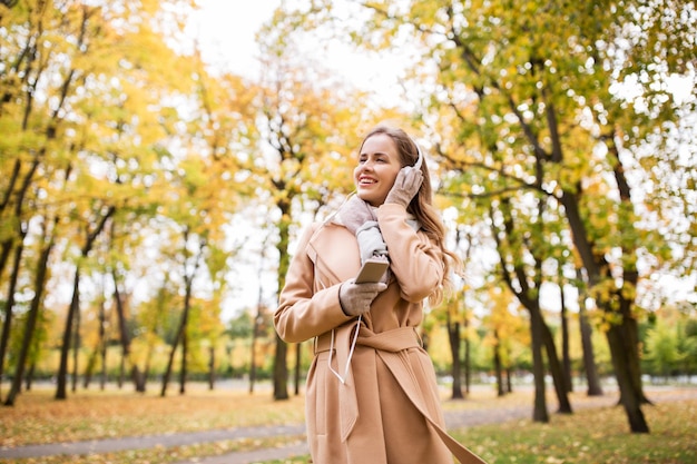 concetto di stagione, tecnologia e persone - bella giovane donna felice con le cuffie che ascolta musica sullo smartphone che cammina nel parco autunnale