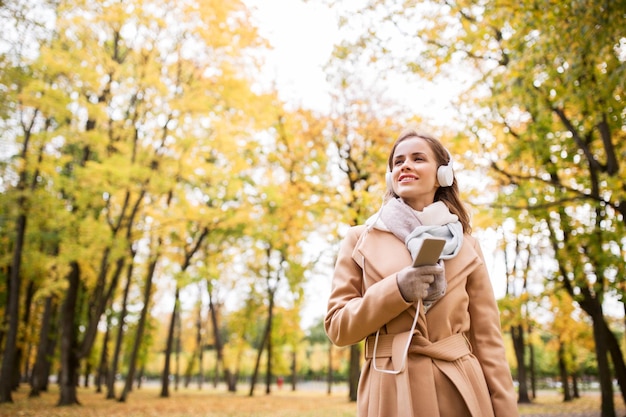 concetto di stagione, tecnologia e persone - bella giovane donna felice con le cuffie che ascolta musica sullo smartphone che cammina nel parco autunnale