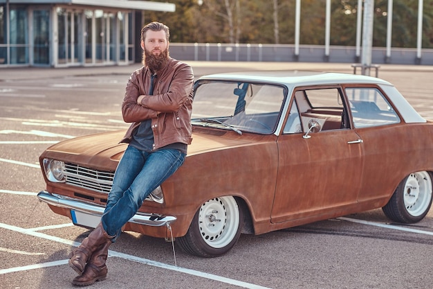 Concetto di stile country. Maschio alla moda che si appoggia sull'auto retrò sintonizzata nel parcheggio della città vicino al grattacielo al tramonto luminoso.