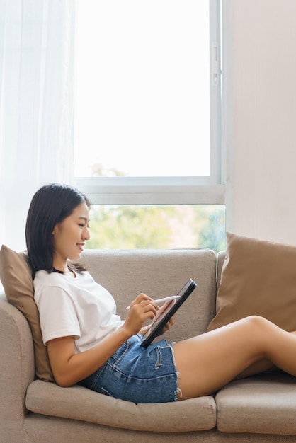 Concetto di stile di vita domestico Giovane donna sdraiata sul divano e che usa il tablet per navigare sui social media a casa
