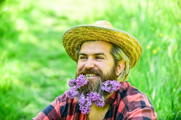 Concetto di stile di vita eco-friendly Uomo rustico con barba faccia felice godersi la vita in ambiente ecologico Hipster con fiori lilla sembra felice Uomo barbuto con lilla in barba sfondo erba verde