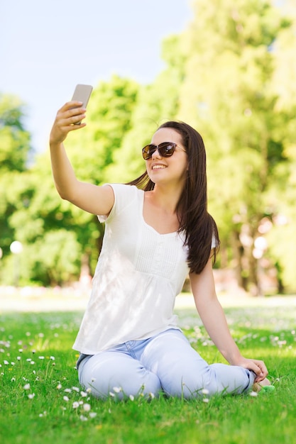 concetto di stile di vita, estate, vacanze, tecnologia e persone - ragazza sorridente con smartphone che fa selfie e si siede sull'erba nel parco