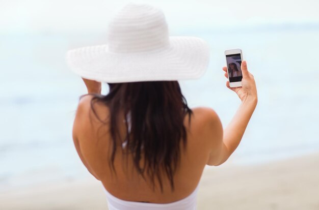 concetto di stile di vita, tempo libero, estate, tecnologia e persone - giovane donna sorridente o adolescente con cappello da sole che si fa selfie con lo smartphone sulla spiaggia