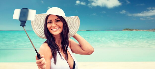 concetto di stile di vita, tempo libero, estate, tecnologia e persone - giovane donna sorridente o ragazza adolescente in cappello da sole che scatta foto con lo smartphone sul selfie stick su sfondo spiaggia tropicale