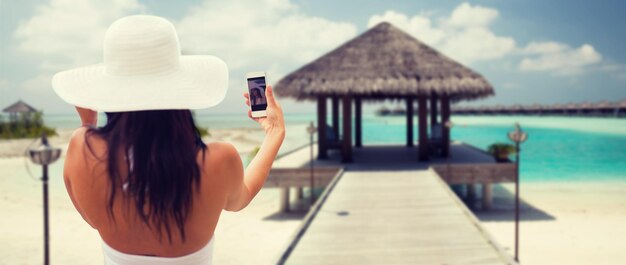 concetto di stile di vita, tempo libero, estate, tecnologia e persone - giovane donna sorridente o ragazza adolescente in cappello da sole che si fa selfie con lo smartphone sopra il bungalow sullo sfondo della spiaggia