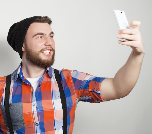 Concetto di stile di vita: un giovane con la barba in camicia che tiene il telefono cellulare e fa una foto di se stesso mentre si trova su uno sfondo grigio