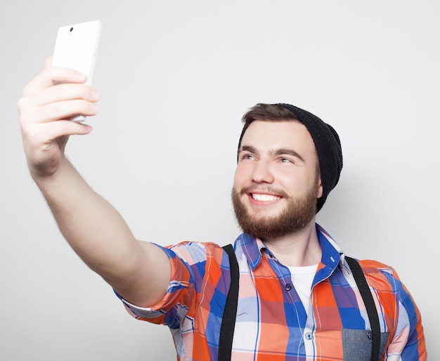 Concetto di stile di vita un giovane uomo con la barba in camicia che tiene il telefono cellulare e fa foto di se stesso mentre si trova in piedi su uno sfondo grigio