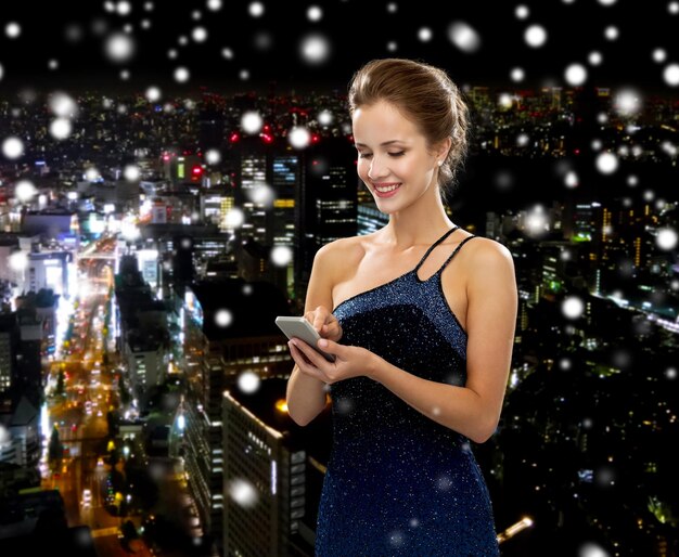 concetto di tecnologia, comunicazione e persone - donna sorridente in abito da sera che tiene smartphone su sfondo di città di notte innevata