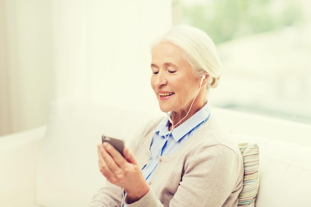 concetto di tecnologia, età e persone - donna anziana felice con smartphone e auricolari che ascolta la musica a casa