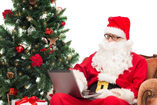 concetto di tecnologia, vacanze e persone - uomo in costume di babbo natale con computer portatile, regali e albero di natale seduto in poltrona