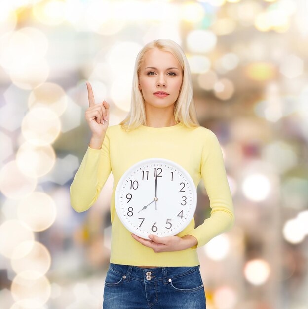 concetto di tempo, istruzione e persone - giovane donna seria con orologio da parete che mostra 8 e dito in alto