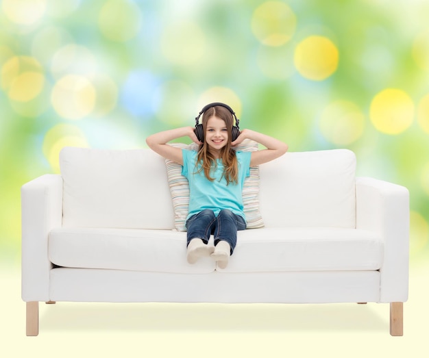 concetto di tempo libero, tecnologia, musica e infanzia - bambina sorridente in cuffia che ascolta musica seduta sul divano su sfondo di luci verdi