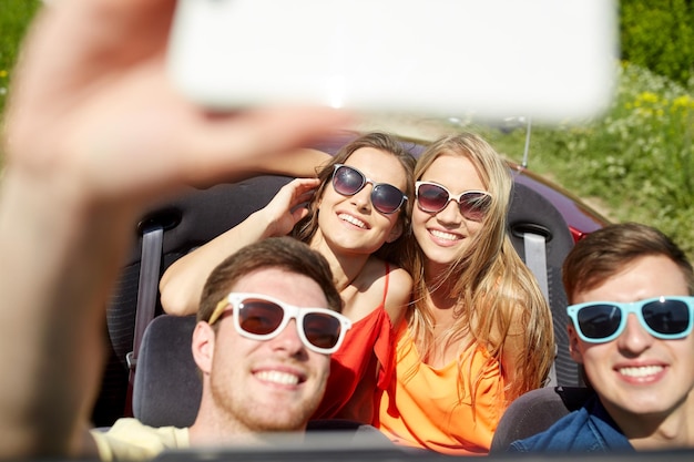 concetto di tempo libero, viaggio su strada, viaggio e persone - amici felici che guidano in cabriolet prendendo selfie con lo smartphone all'aperto