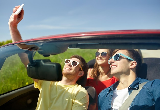 concetto di tempo libero, viaggio su strada, viaggio e persone - amici felici che guidano in cabriolet prendendo selfie con lo smartphone all'aperto