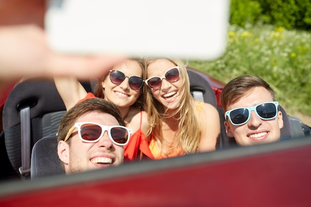 concetto di tempo libero, viaggio su strada, viaggio e persone - amici felici che guidano in cabriolet prendendo selfie con lo smartphone all'aperto