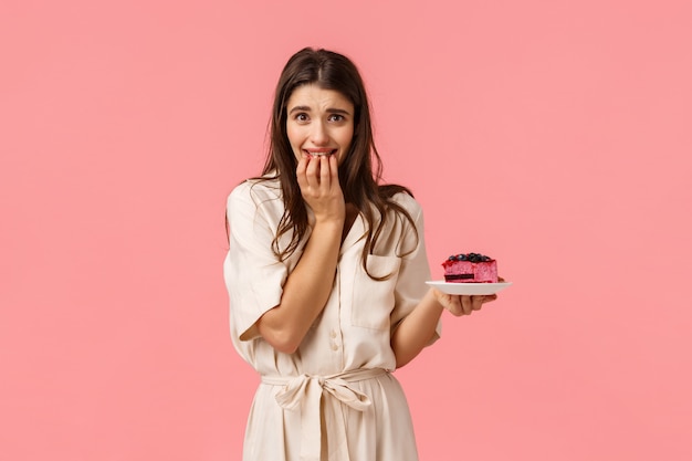 Concetto di tentazione, dieta e dessert. Seducente giovane ragazza bruna sportiva femminile in abito, cercando di perdere peso per l'estate, tenendo in mano un pezzo di torta e mordicchiando le dita vuoi mangiare ma non posso, muro rosa