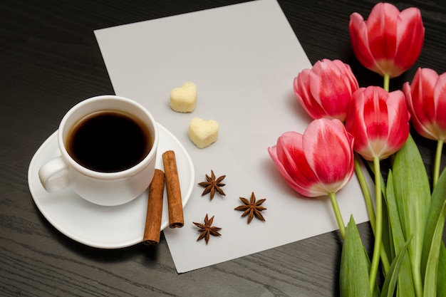 Concetto di vacanza. Bouquet di tulipani rosa, una tazza di caffè, zucchero a forma di cuore, cannella, anice stellato e foglio di carta