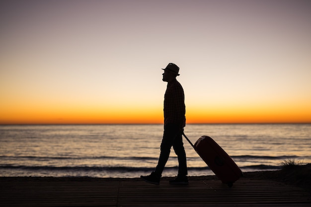 Concetto di vacanza, estate e viaggio - silhouette di giovane uomo con la valigia al tramonto vicino al mare.