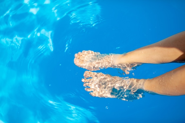 Concetto di vacanza estiva. Le gambe di una giovane donna sullo sfondo dell'acqua blu della piscina. Nuotando nella piscina. Relax, abbronzatura e allegria.