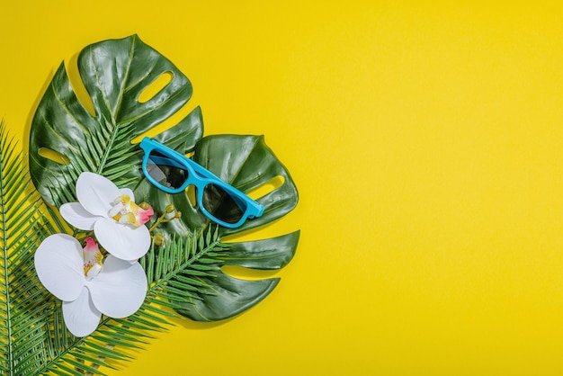 Concetto di vacanza estiva Occhiali da sole decorazioni marine palme e foglie di monstera fiore di orchidea Sfondo giallo brillante luce dura ombra scura vista dall'alto piatta