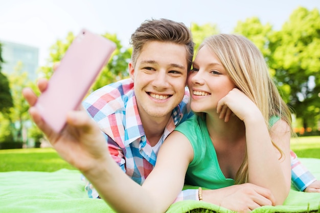 concetto di vacanza, vacanze, tecnologia e amicizia - coppia sorridente sdraiata su una coperta e fare selfie con lo smartphone nel parco