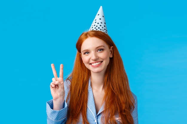 Concetto di vacanze, celebrazione e congratulazioni. Donna europea rossa allegra con cappello b-day che mostra il segno di pace, indossa indumenti da notte, festeggia il compleanno, sorridendo su sfondo blu