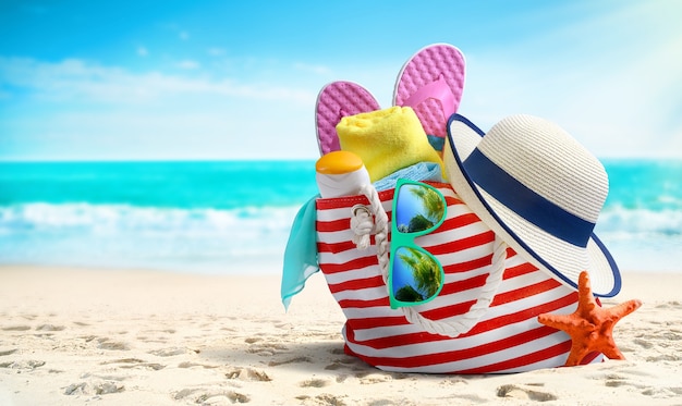 Concetto di vacanze estive. Accessori da spiaggia - borsa da spiaggia, cappello di paglia, infradito e occhiali da sole sulla spiaggia sabbiosa e mare azzurro sullo sfondo