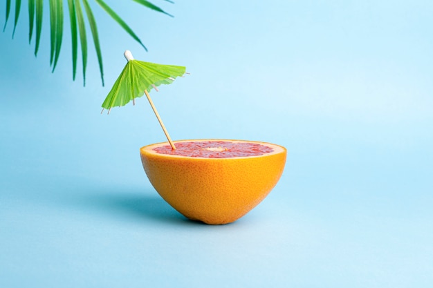 Concetto di vacanze estive. Pompelmo con un ombrello da cocktail su uno sfondo colorato. Tropici, sole, spiaggia, vitamine, frutta, estate e buon umore