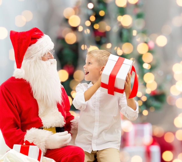 concetto di vacanze, infanzia e persone - ragazzino sorridente con babbo natale e regali su sfondo di luci albero di natale