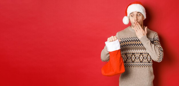 Concetto di vacanze invernali Capodanno e celebrazione Uomo adulto sorpreso che tiene calza di natale con regali all'interno guardando stupito dalla borsa in piedi su sfondo rosso in cappello da Babbo Natale