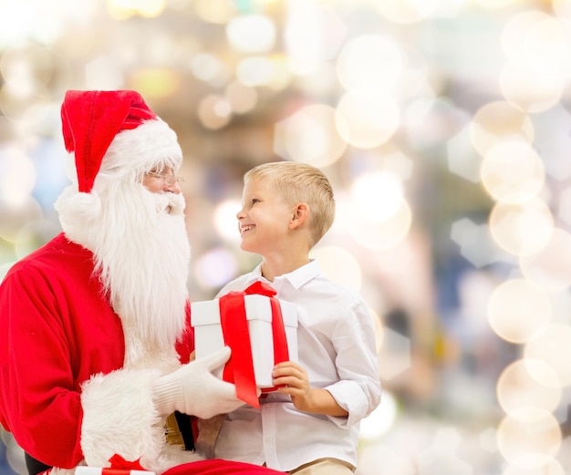 concetto di vacanze, natale, infanzia e persone - ragazzino sorridente con babbo natale e regali su sfondo di luci