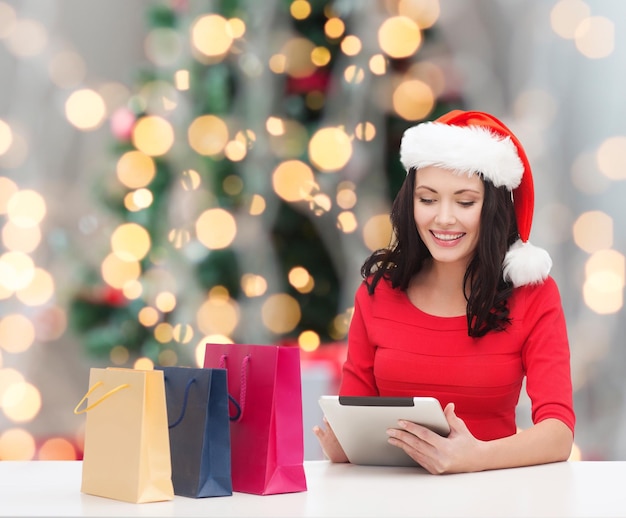 concetto di vacanze, tecnologia e persone - donna sorridente in cappello di Babbo Natale con borse della spesa e computer tablet pc sopra l'albero di Natale con sfondo di luci
