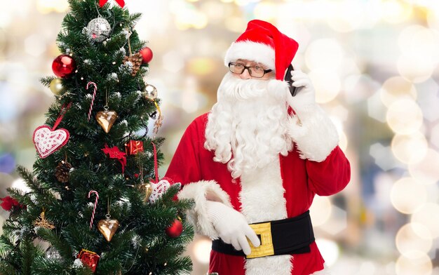 concetto di vacanze, tecnologia e persone - uomo in costume di babbo natale con smartphone e albero di natale su sfondo di luci