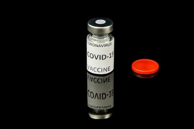 Concetto di vaccino. Bottiglia di vetro piccola con tappo argento e rosso e firma del vaccino contro il Coronavirus Covid 19 su sfondo nero lucido. Avvicinamento. Messa a fuoco selettiva. Copia spazio