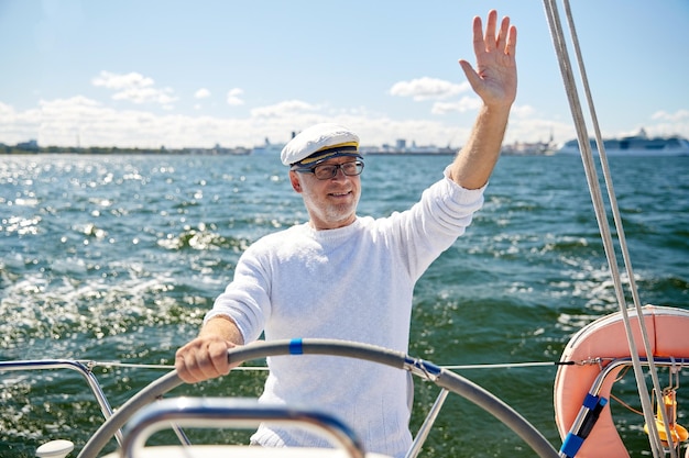 concetto di vela, età, turismo, viaggi e persone - uomo anziano felice con cappello da capitano sul volante e barca a vela o yacht che galleggia nel mare