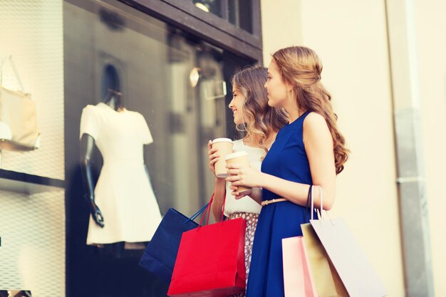 concetto di vendita, consumismo e persone - giovani donne felici con borse della spesa e tazze di carta da caffè che guardano la vetrina all'aperto