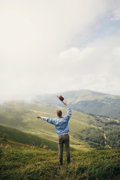 Concetto di viaggio e voglia di viaggiare uomo elegante viaggiatore con cappello in piedi in cima a montagne soleggiate in nuvole spazio per testo ragazzo hipster che viaggia incredibile momento atmosferico