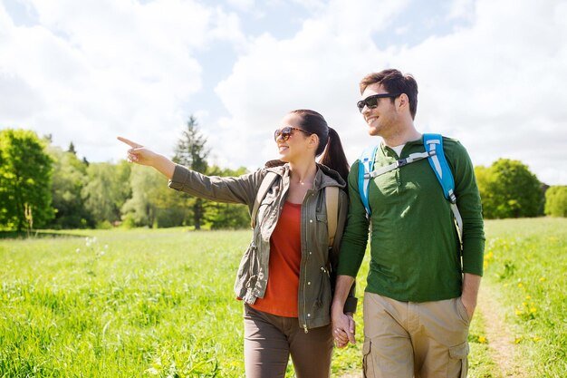 concetto di viaggio, escursionismo, zaino in spalla, turismo e persone - coppia felice con zaini che camminano lungo la strada di campagna all'aperto e puntano il dito verso qualcosa