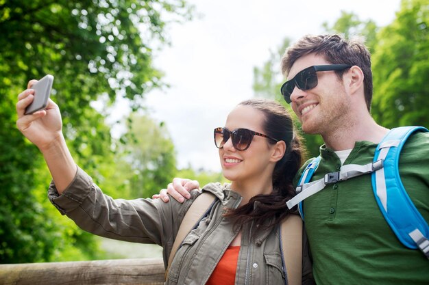 concetto di viaggio, escursionismo, zaino in spalla, turismo e persone - coppia sorridente con zaini che prendono selfie con lo smartphone in natura