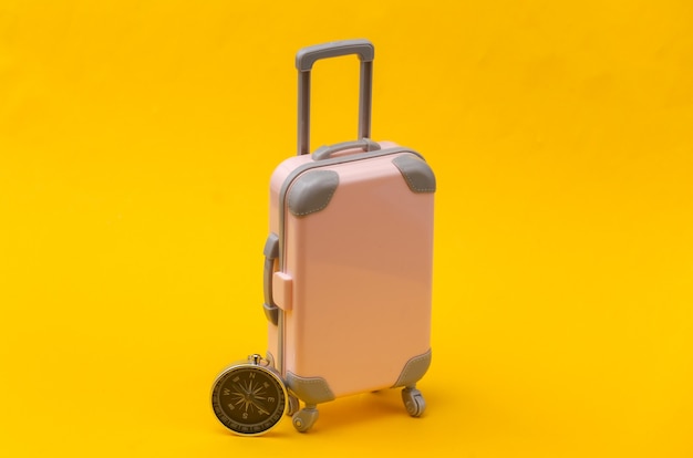 Concetto di viaggio o viaggio. Mini valigia da viaggio in plastica rosa e bussola su sfondo giallo. Stile minimal