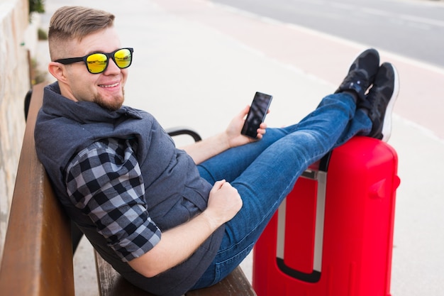 Concetto di viaggio, turismo e persone - bell'uomo si siede e mette i piedi sulla valigia, guardando qualcosa nel suo cellulare.